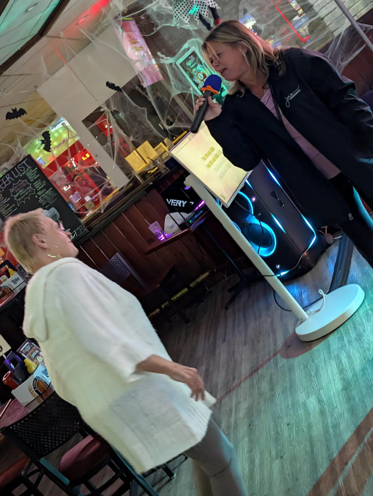 Two guests singing beside the white Samsung lyrics screen on its rolling stand at Coach’s Corner